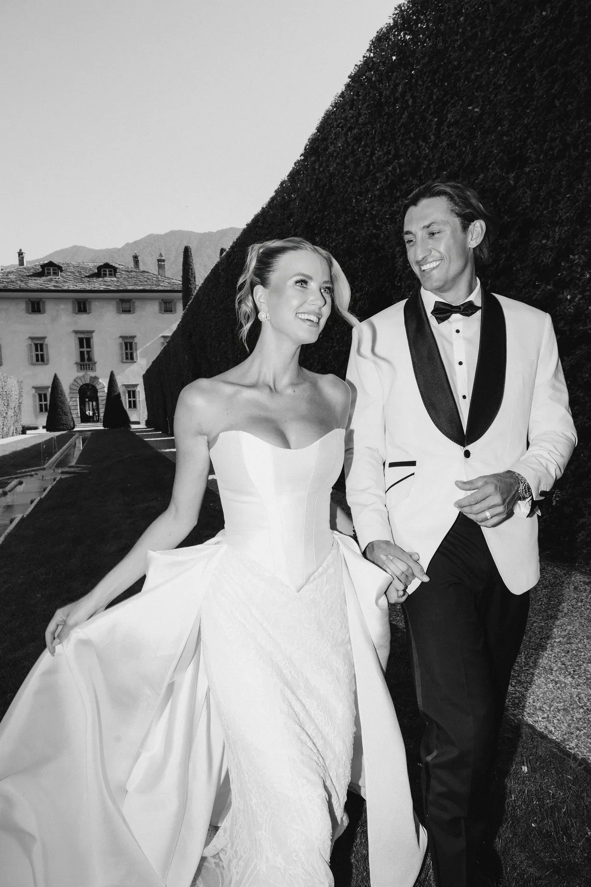 The couple walking a gravel path between tall clipped hedges, the villa behind them