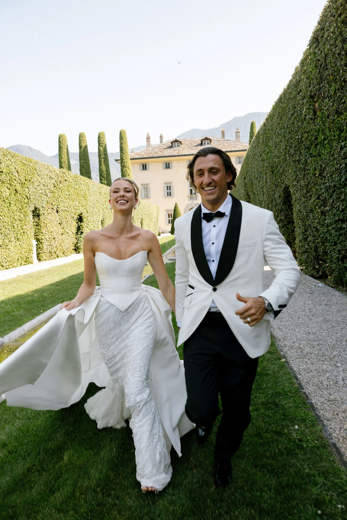 Bride and groom running up the lawn between hedges, the villa and cypresses beyond