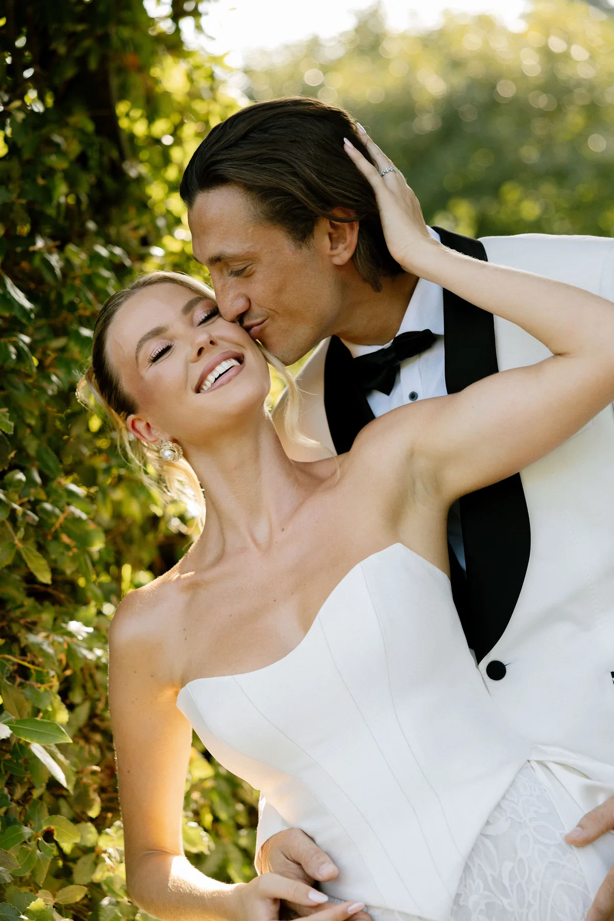 The groom kissing the bride's cheek in low sun, leaves lit behind them