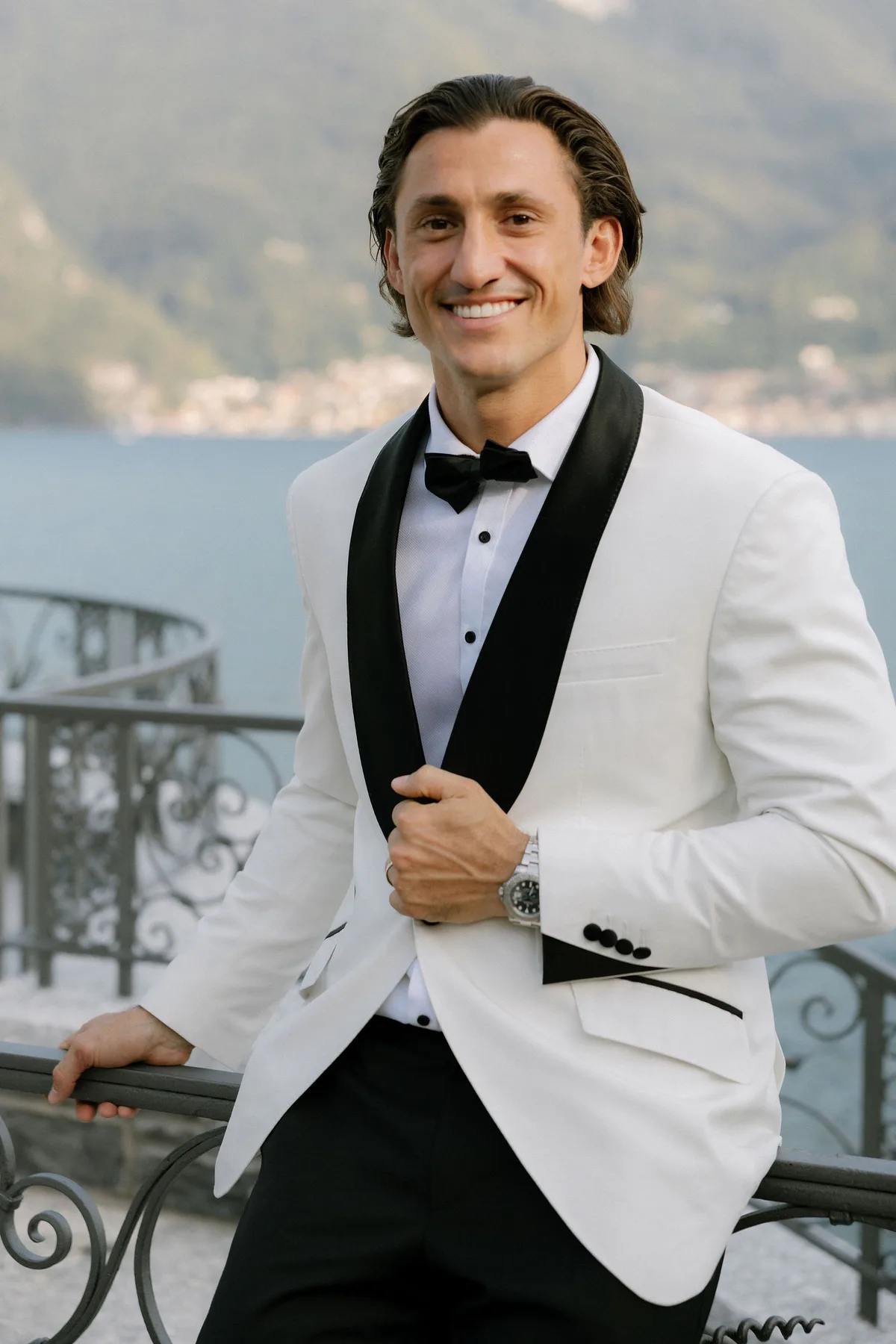 The groom in a white dinner jacket at an iron balustrade above the lake