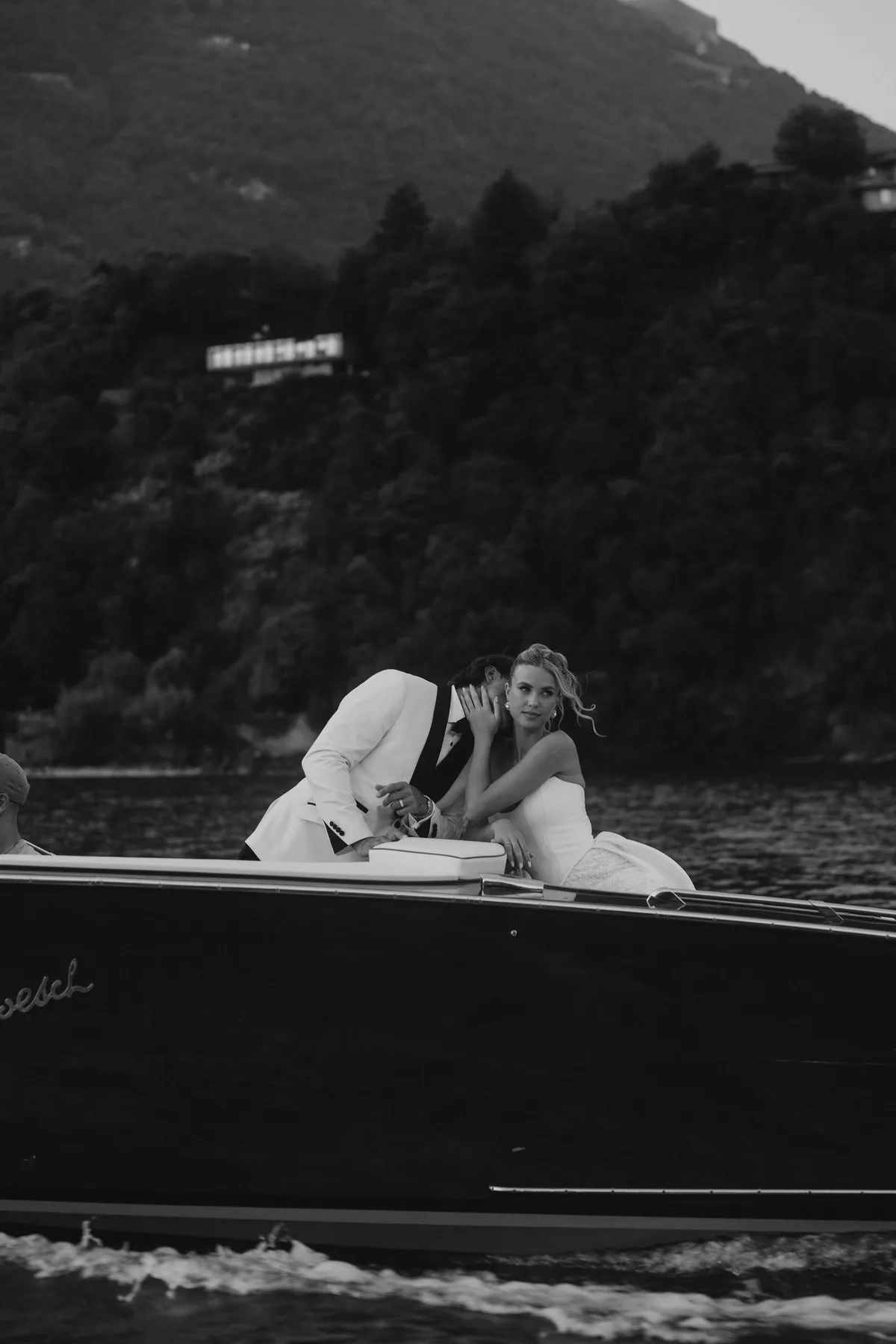The couple in a wooden motor launch crossing the lake, a wooded shore behind