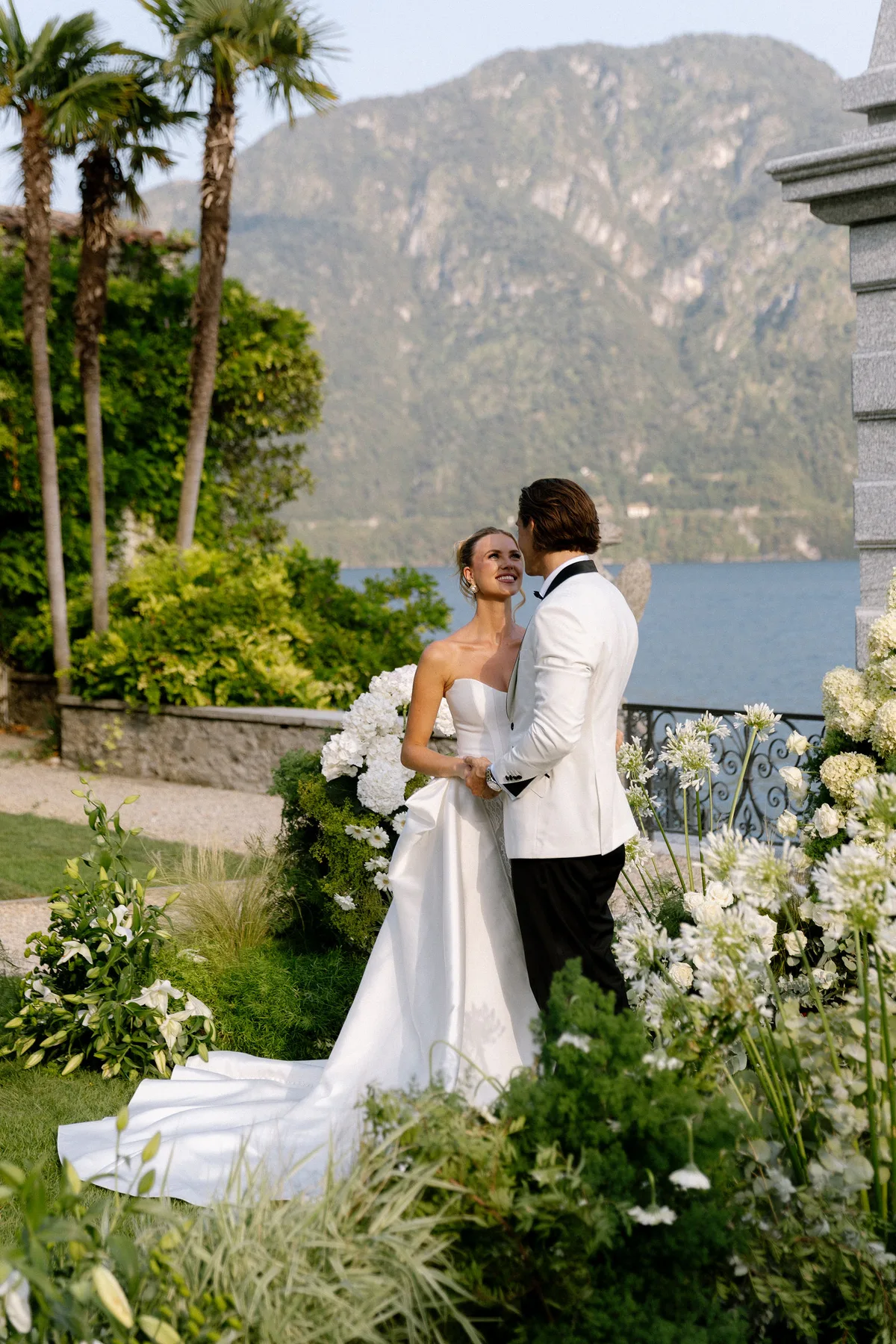 Bride and groom among white flowers on a terrace, palms and the lake beyond