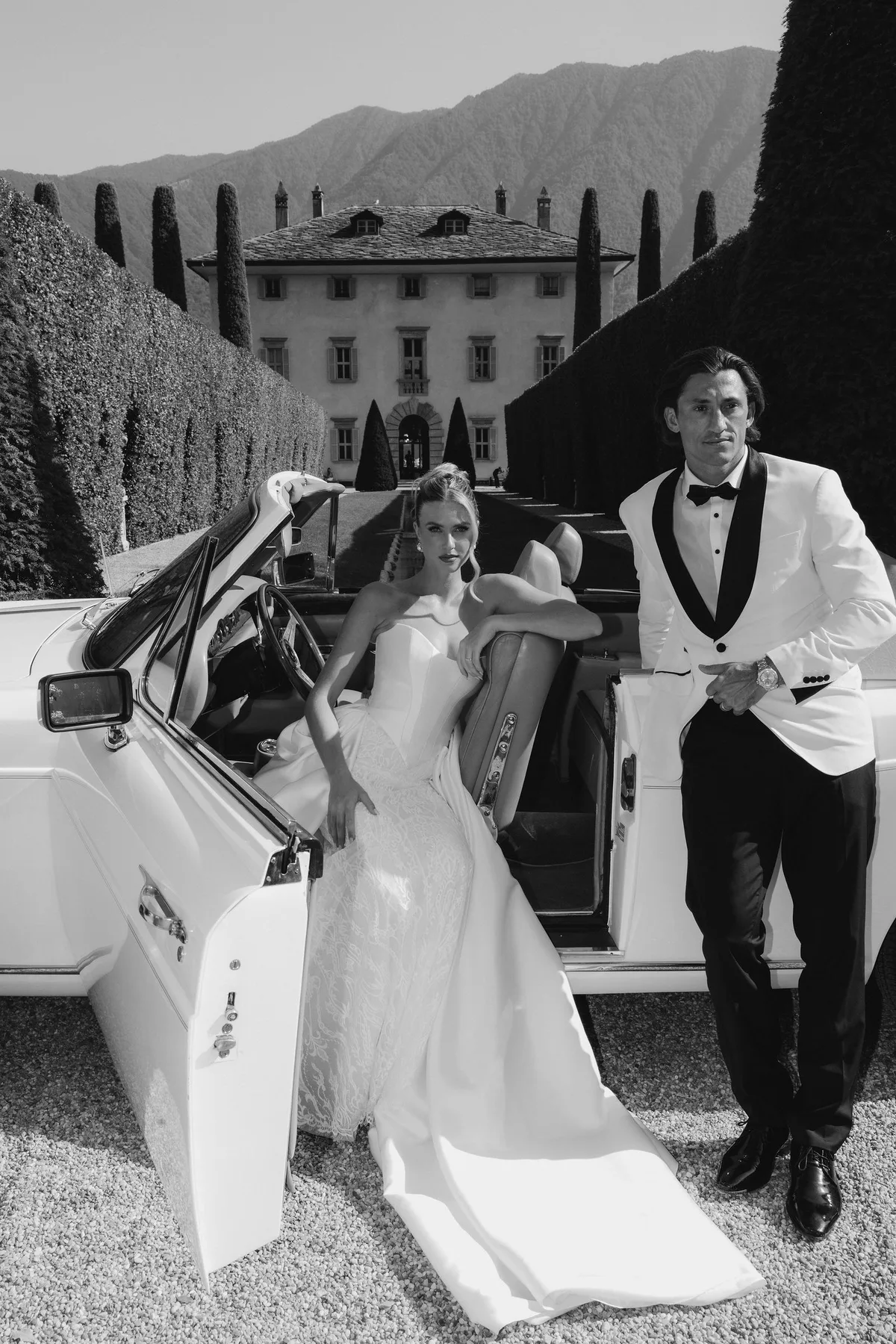 The couple beside an open car on the gravel, the villa and hedges behind