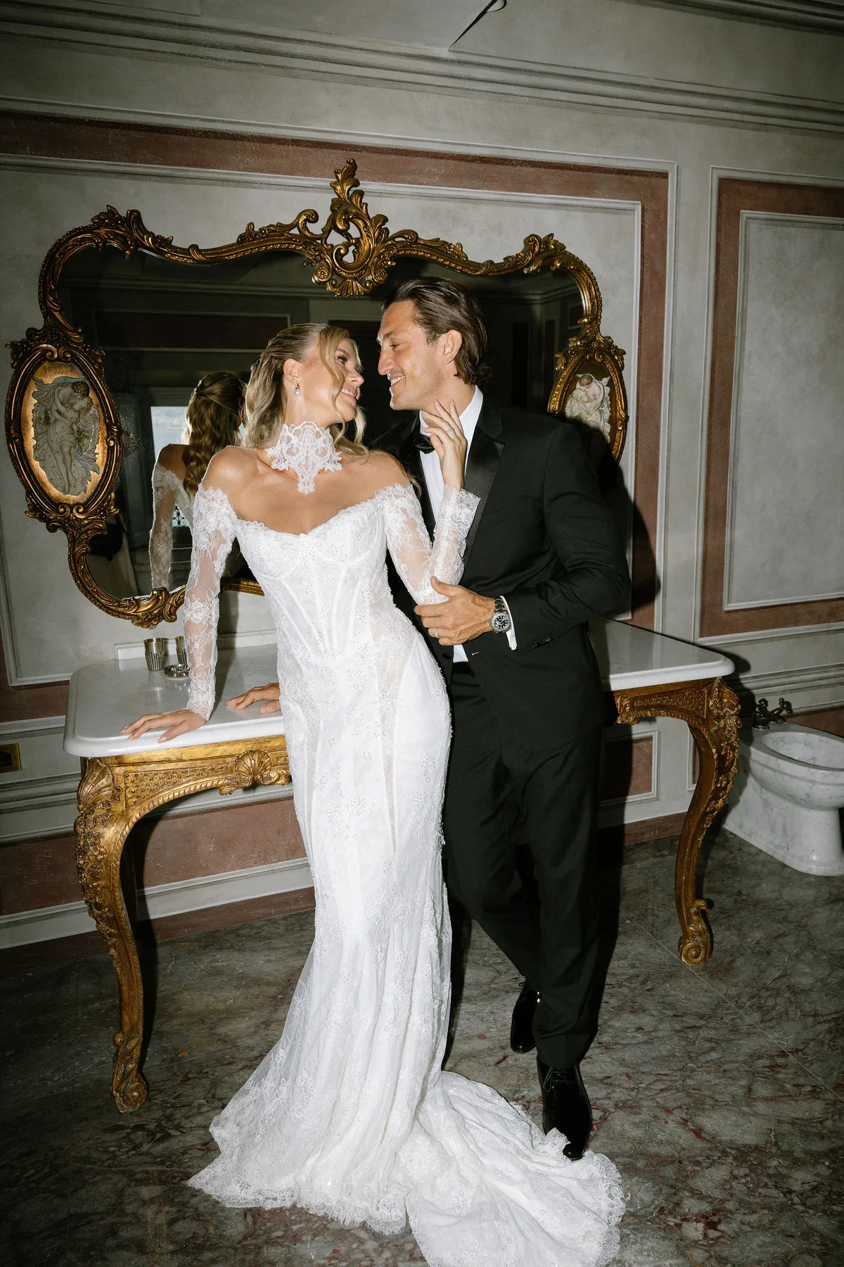 The couple by a gilt console and mirror, the bride's lace train across the marble floor