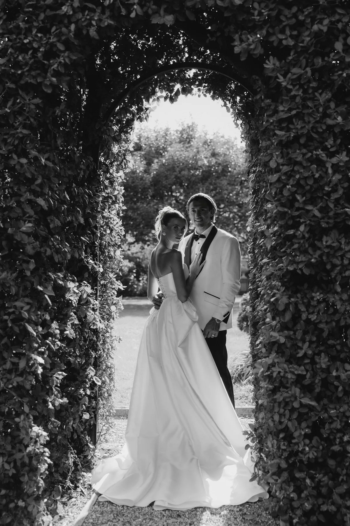 Bride and groom framed by an archway cut through a hedge, the garden beyond