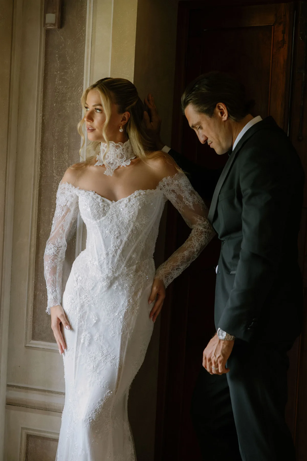 The bride against a doorframe in a lace gown, the groom close beside her