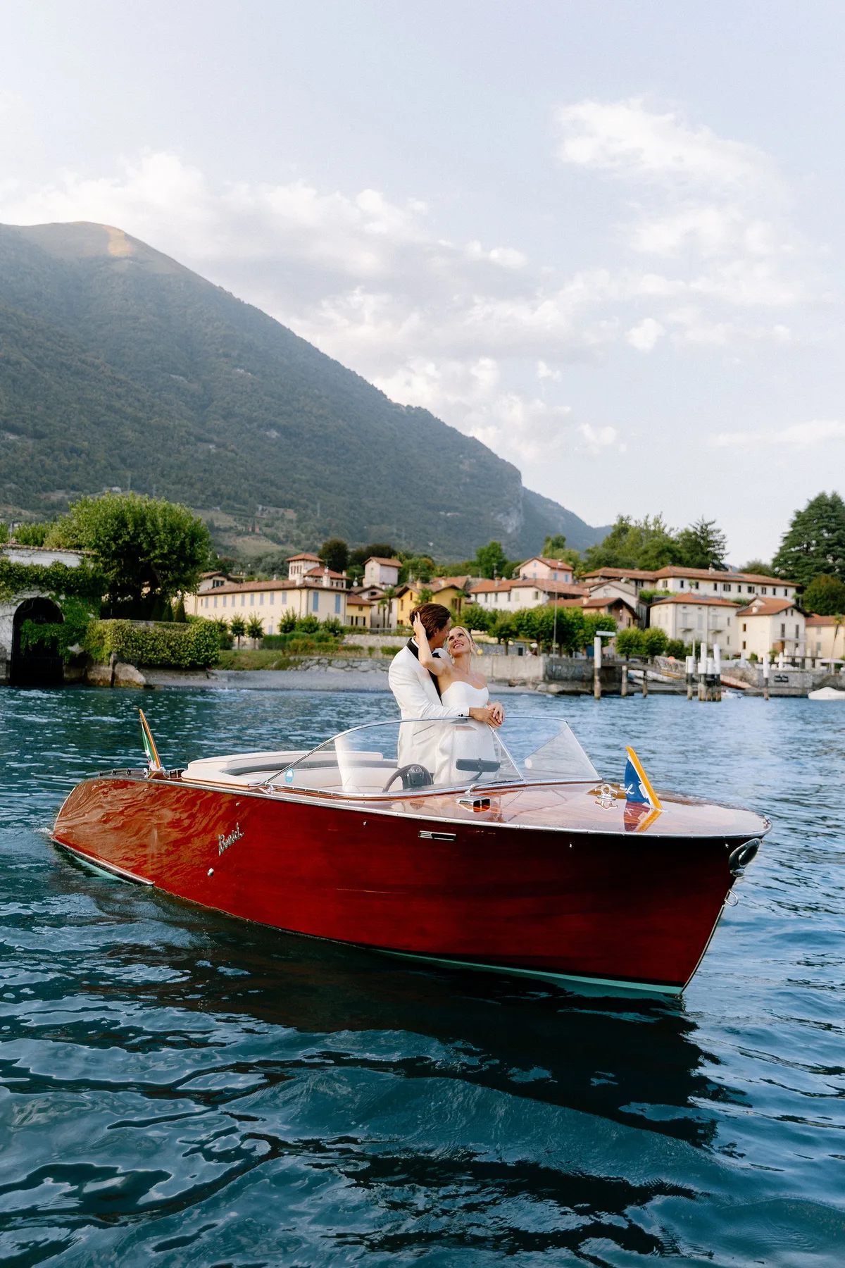 A wooden motor boat on the open lake, the couple aboard, a village on the shore