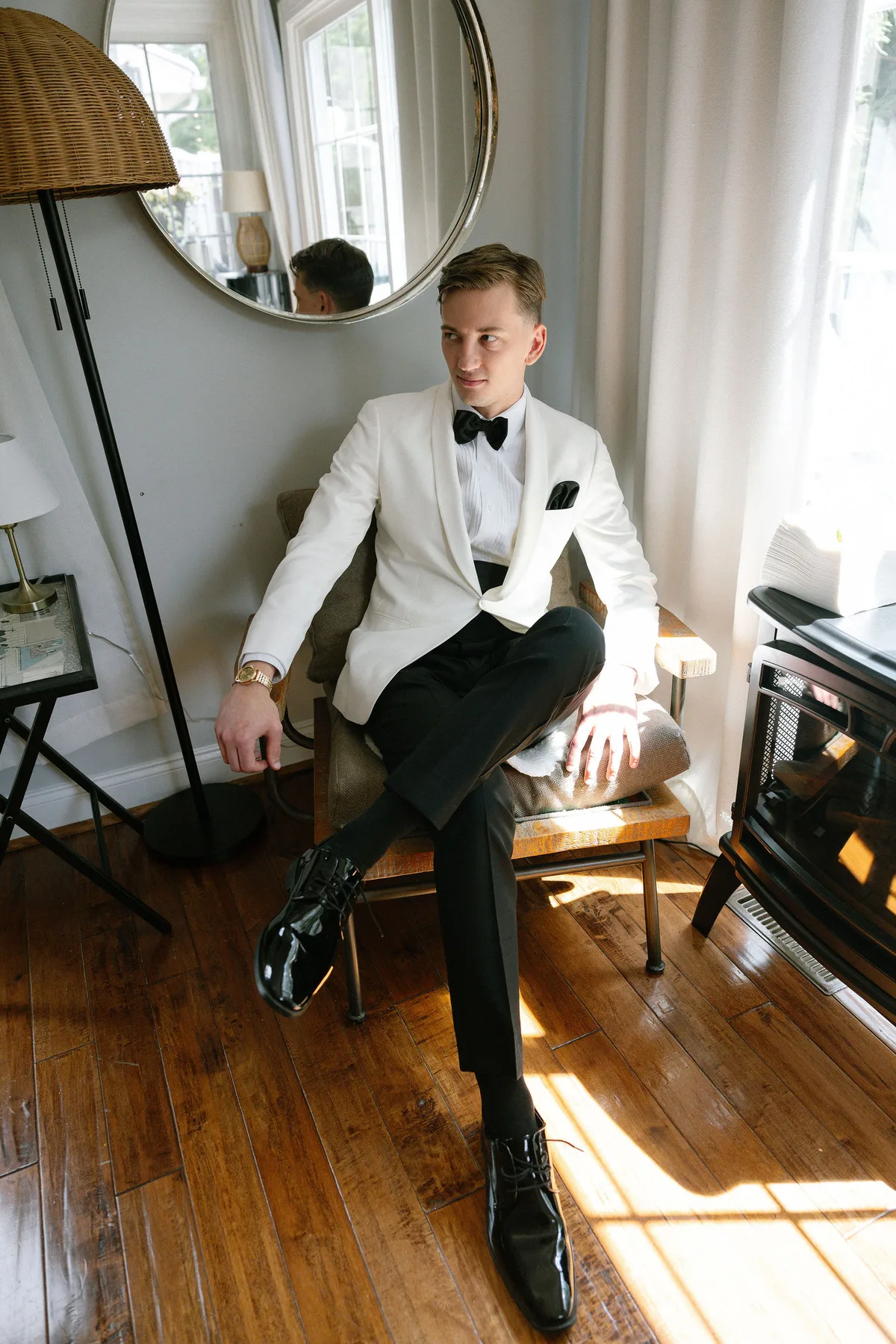 The groom in a white dinner jacket seated by a window, a round mirror behind him