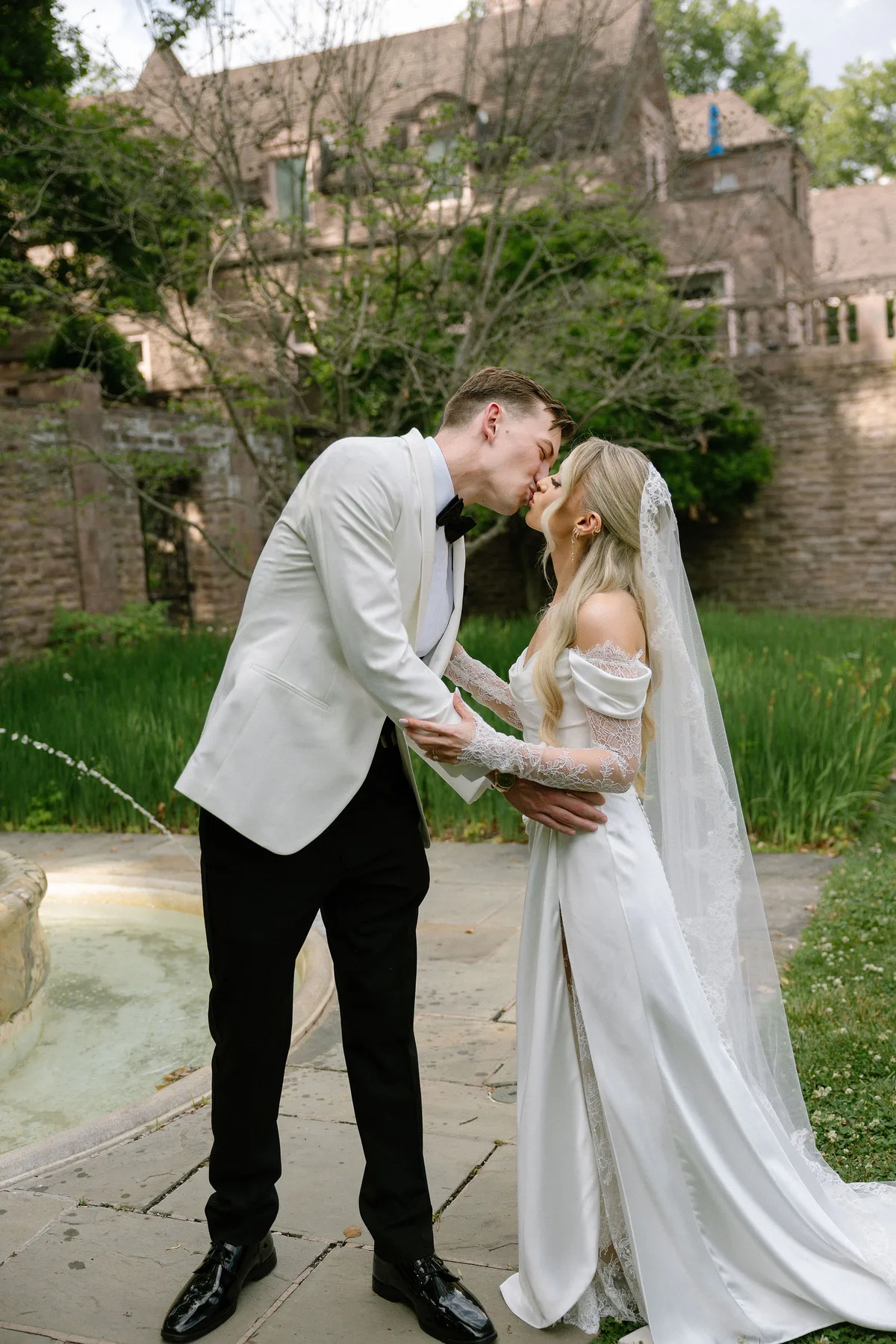 Bride and groom kissing beside a stone fountain, the manor house behind them