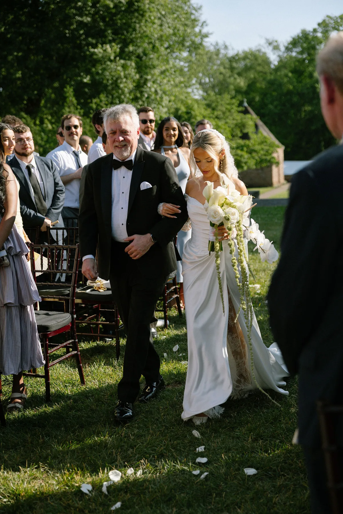 The bride walking down the grass aisle on her father's arm, guests standing