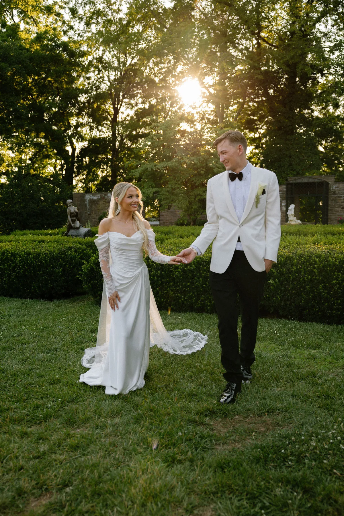 The couple hand in hand on the lawn, low sun through the trees behind them