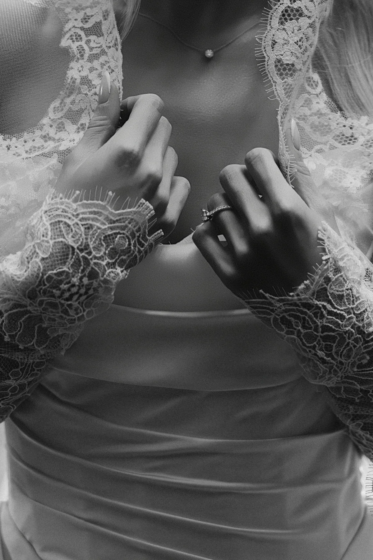 The bride's hands at the neckline of her satin gown, lace sleeves