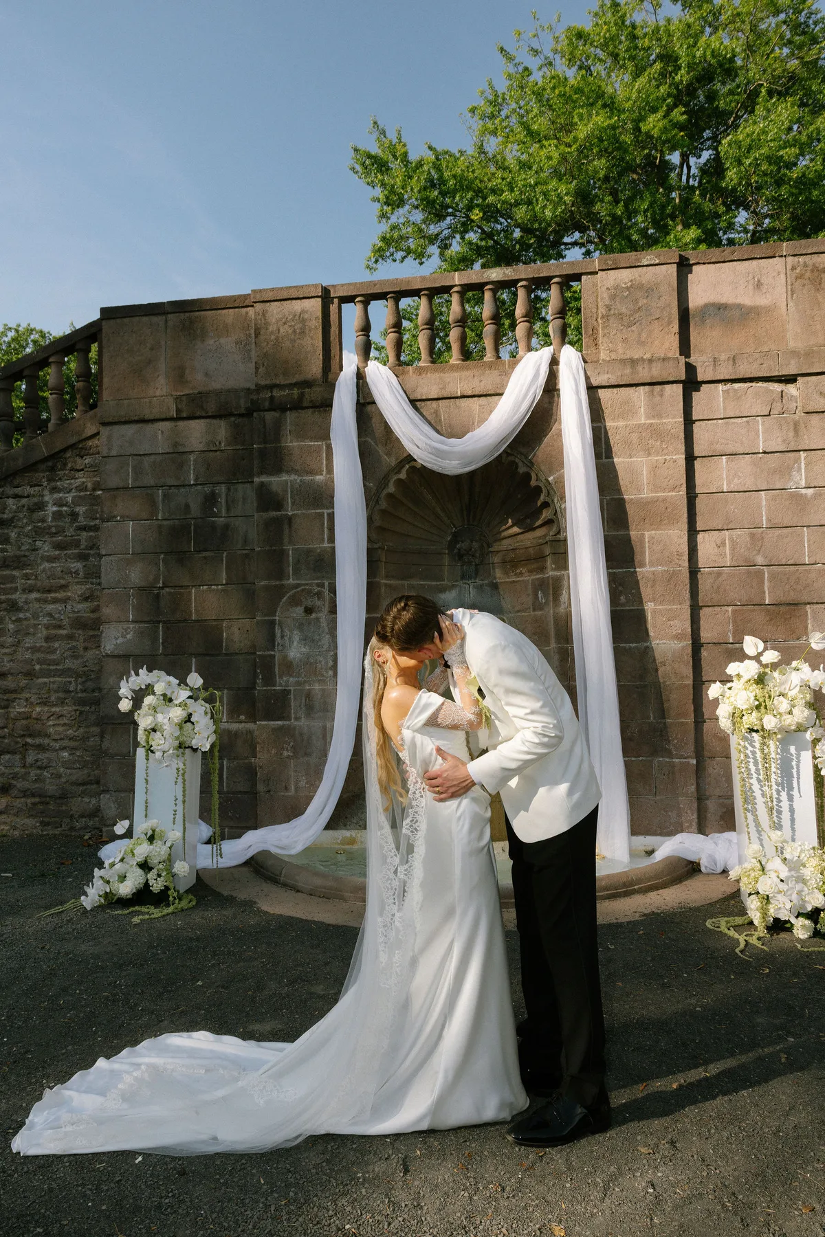 Bride and groom kissing under a draped arch before a stone fountain