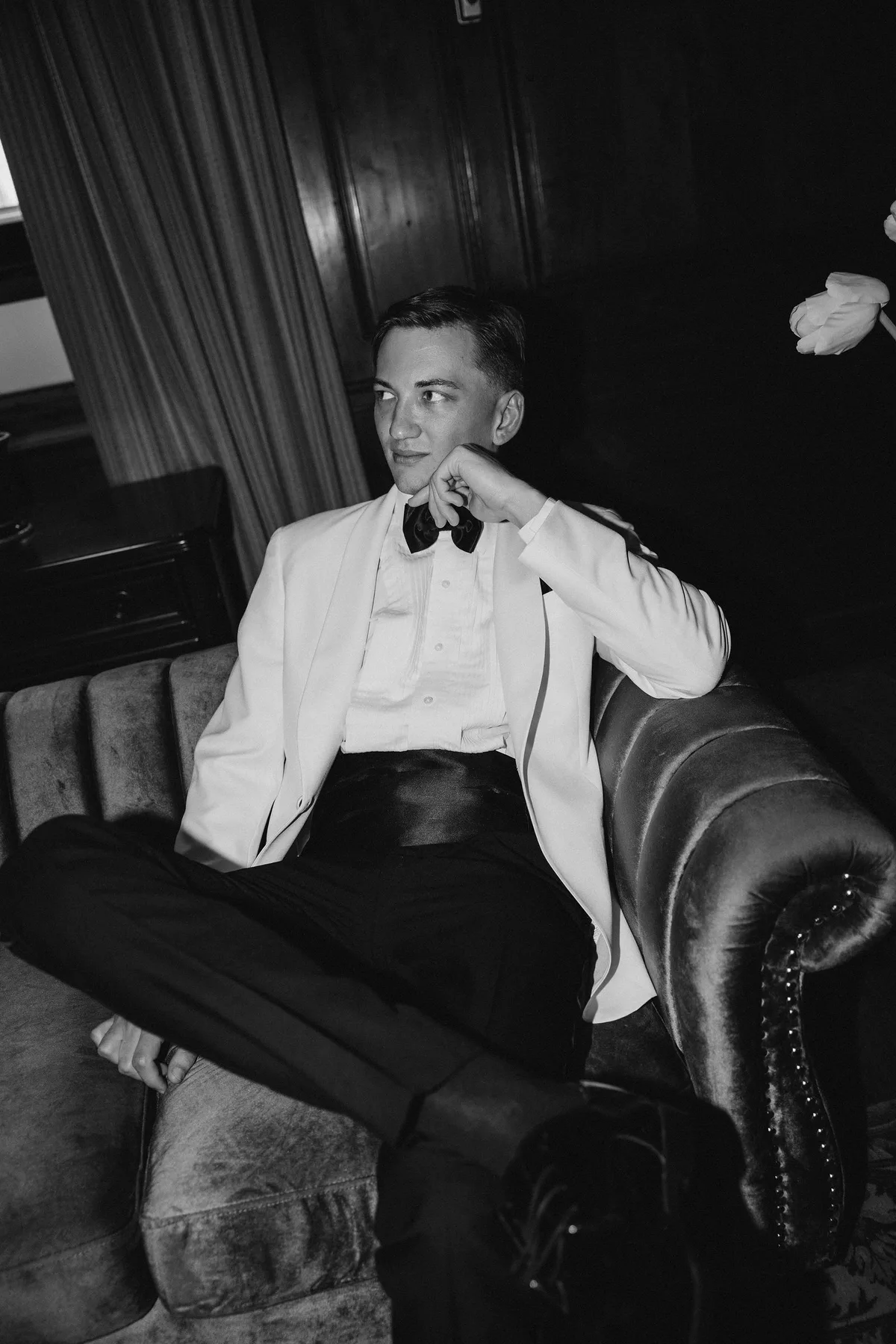 The groom sitting back on a velvet sofa in a dark room