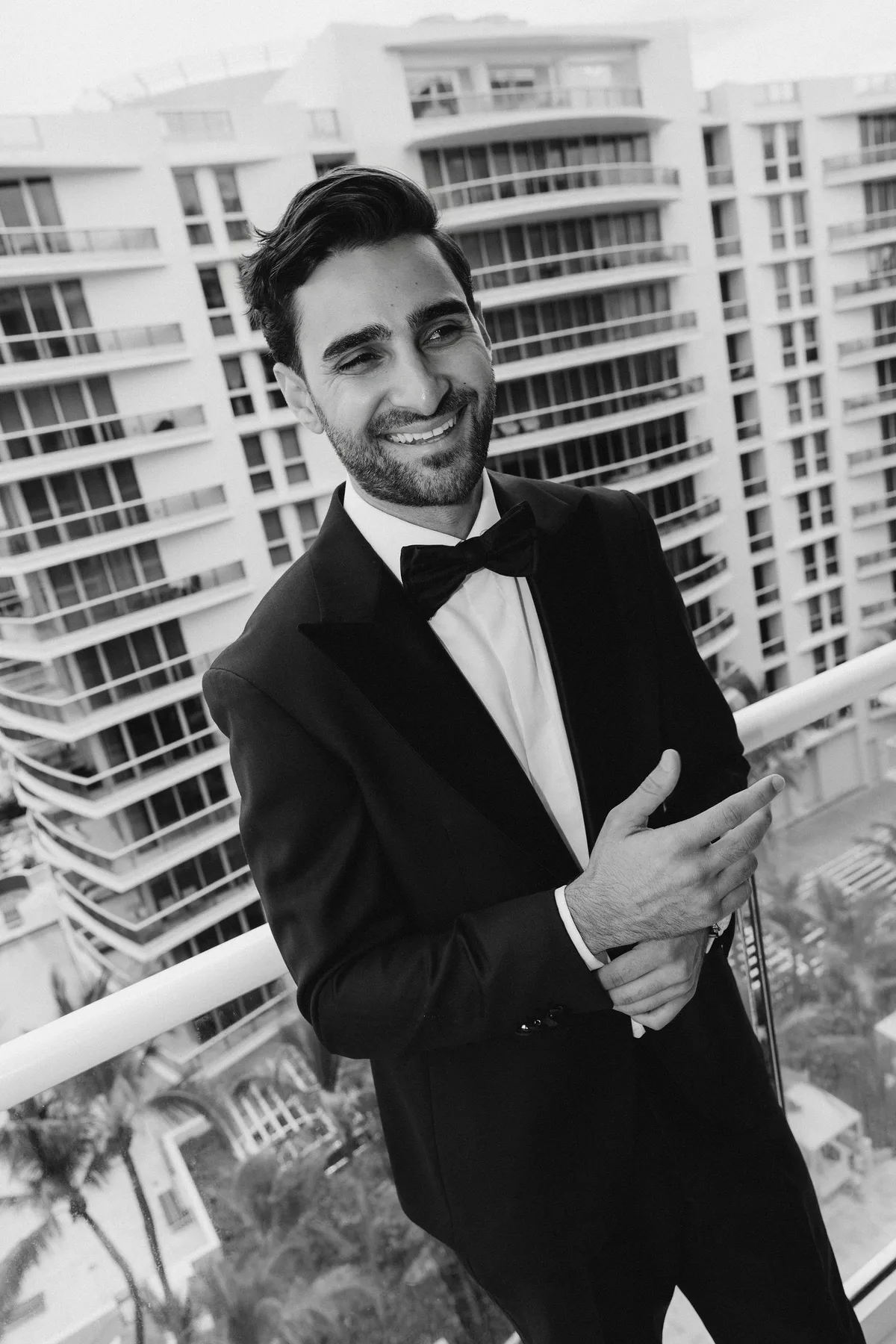The groom in a black tuxedo on a balcony, a white high-rise and palms behind