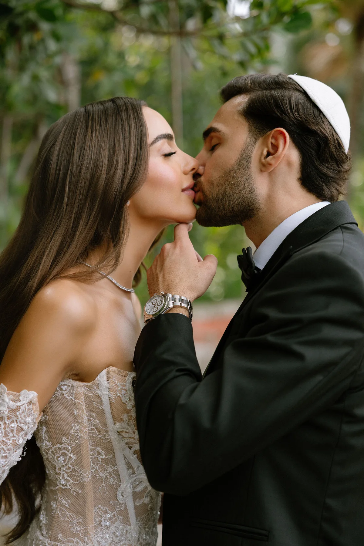 The groom kissing the bride, his hand at her chin, trees behind