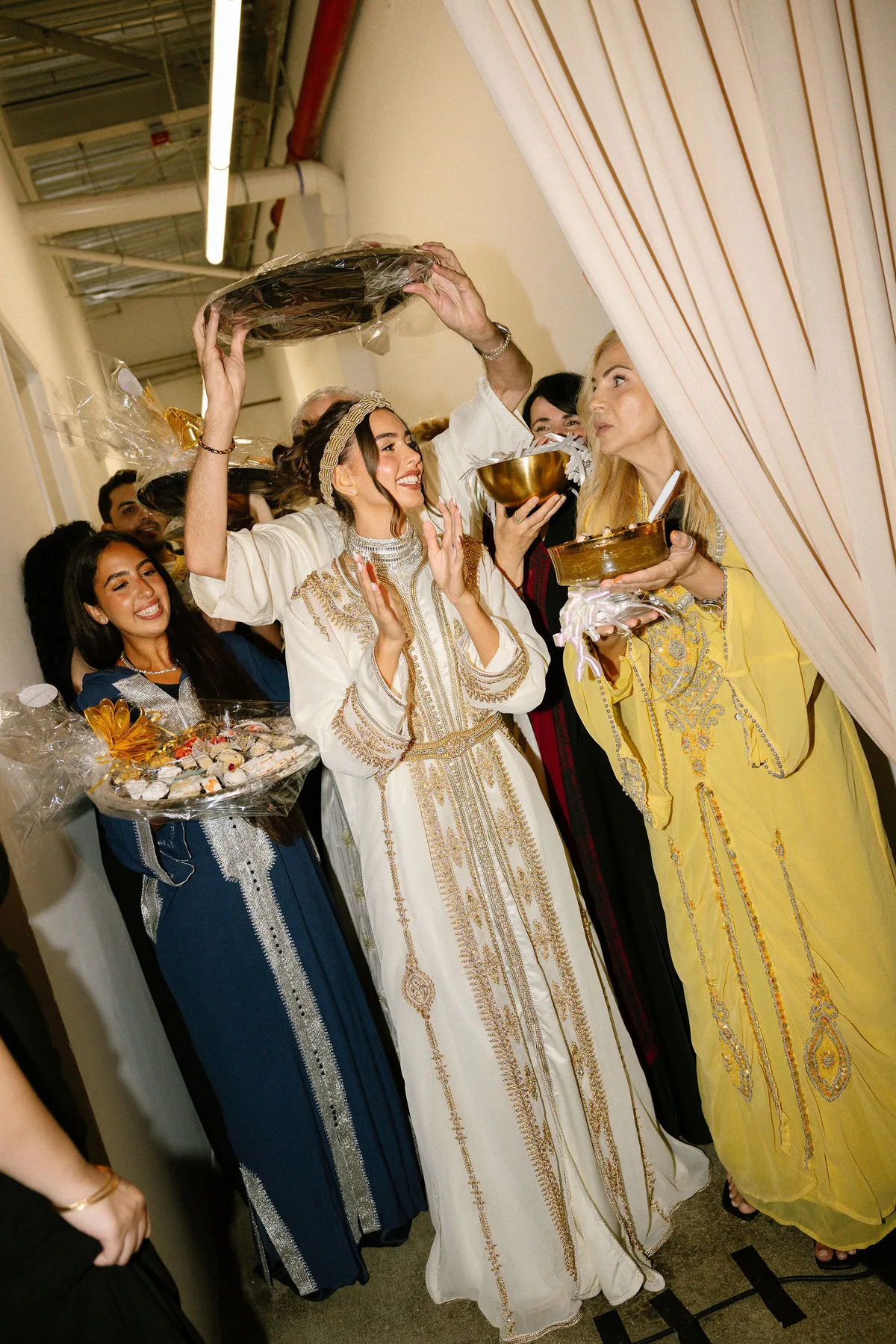 The bride in an embroidered kaftan under a tray held overhead, women in kaftans around her
