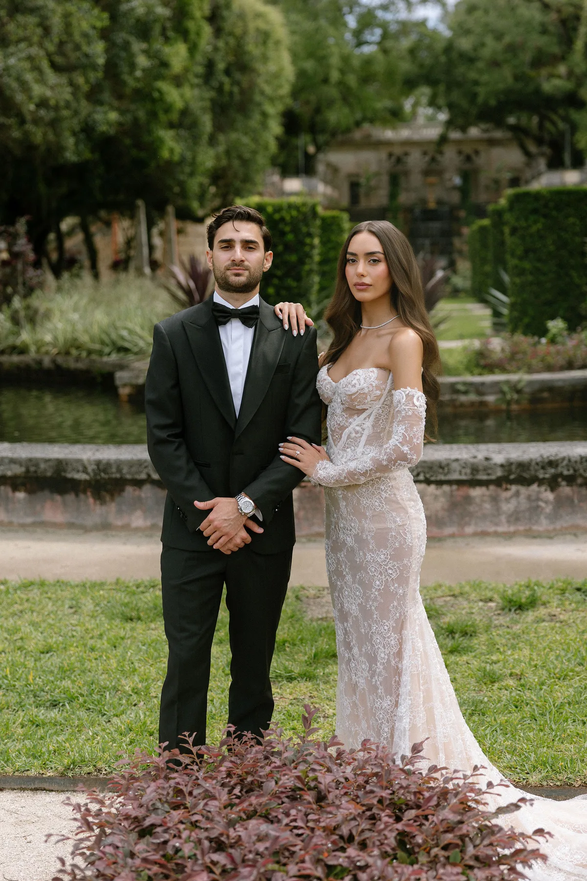 The couple standing by a formal pool, the house behind them