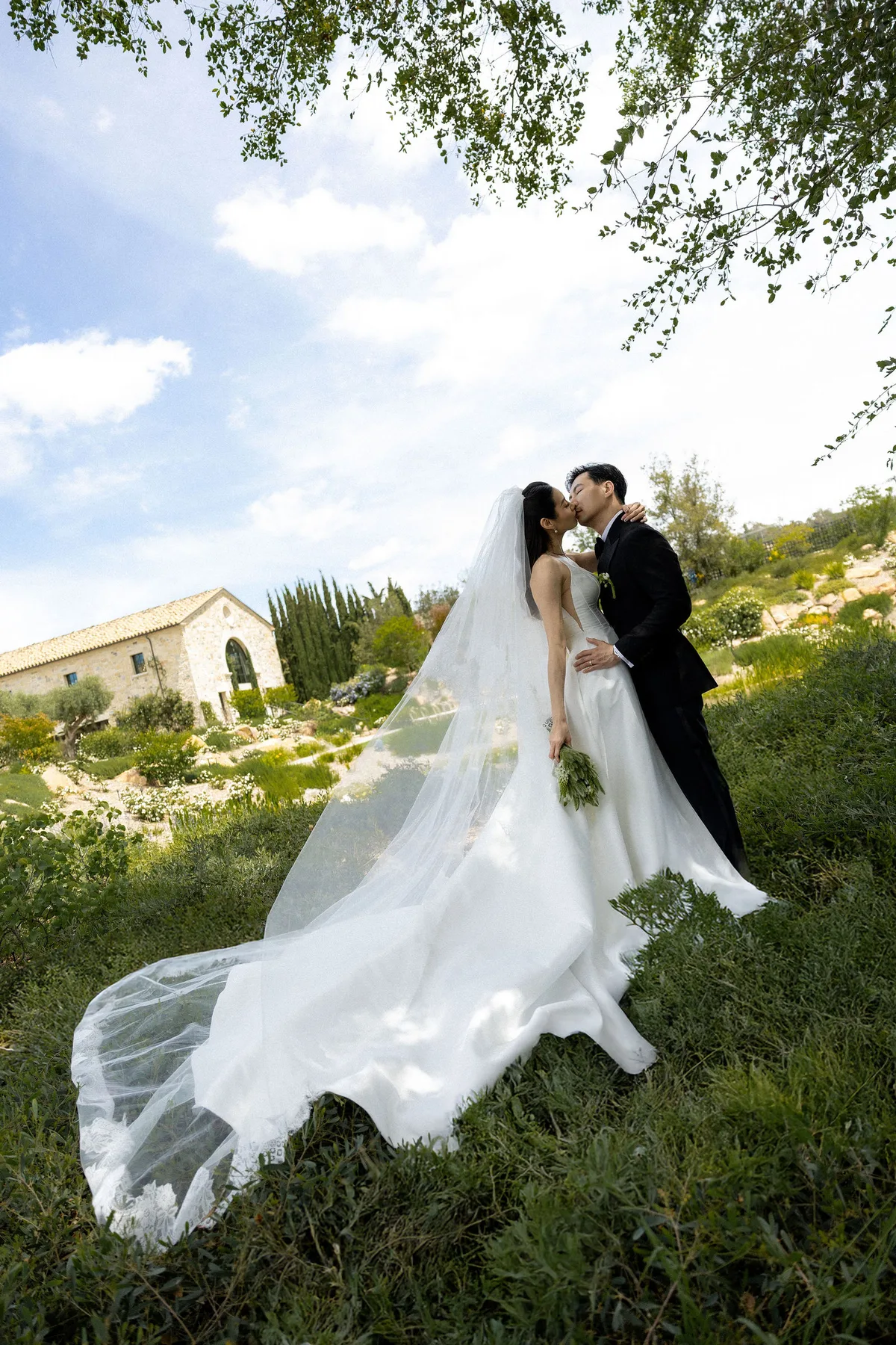 The couple kissing on a slope above the winery, her veil blown out across the grass