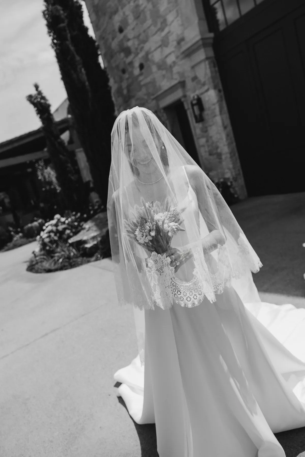 The bride under her veil with a bouquet, cypresses and the stone building behind