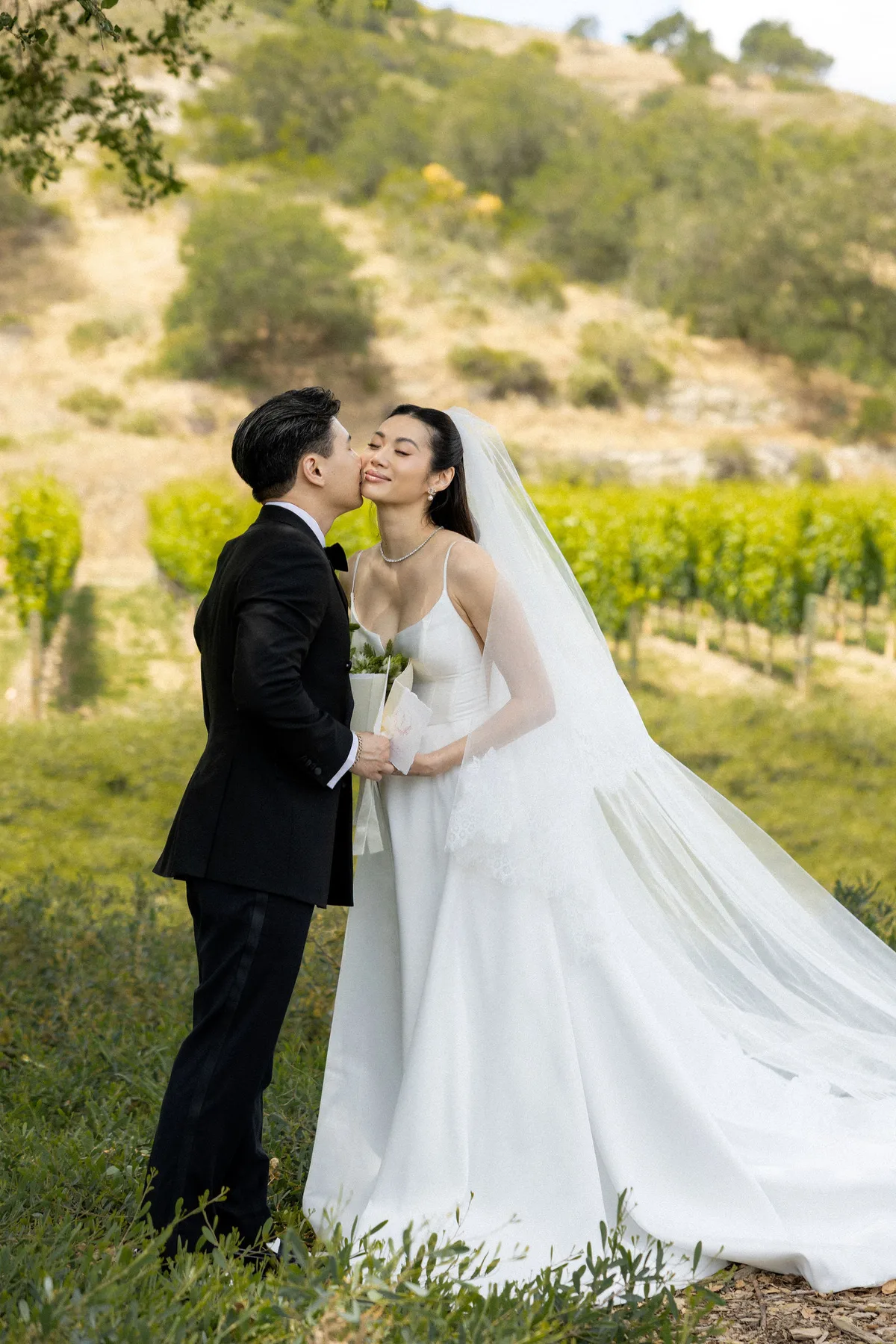 The groom kissing the bride's cheek on the grass, rows of vines behind