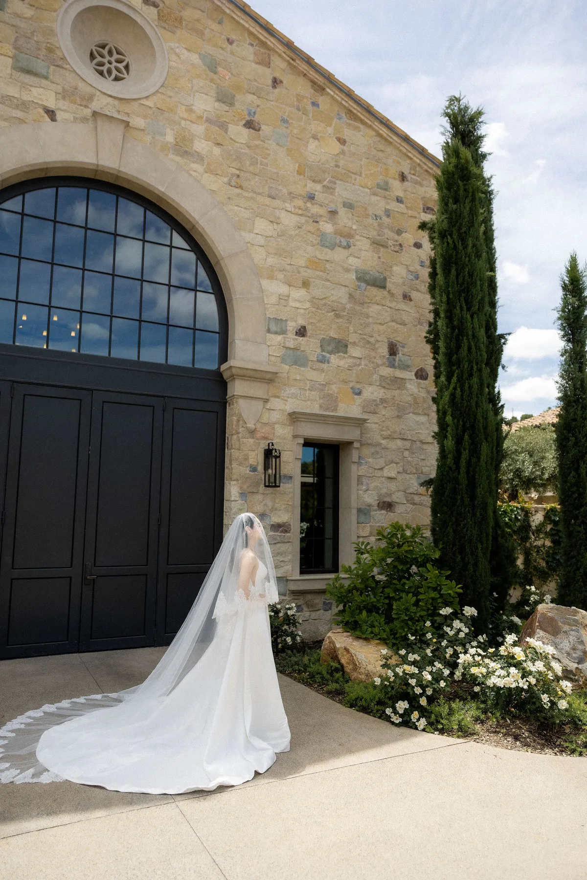 The bride small before the arched doors of the stone winery, cypresses beside her