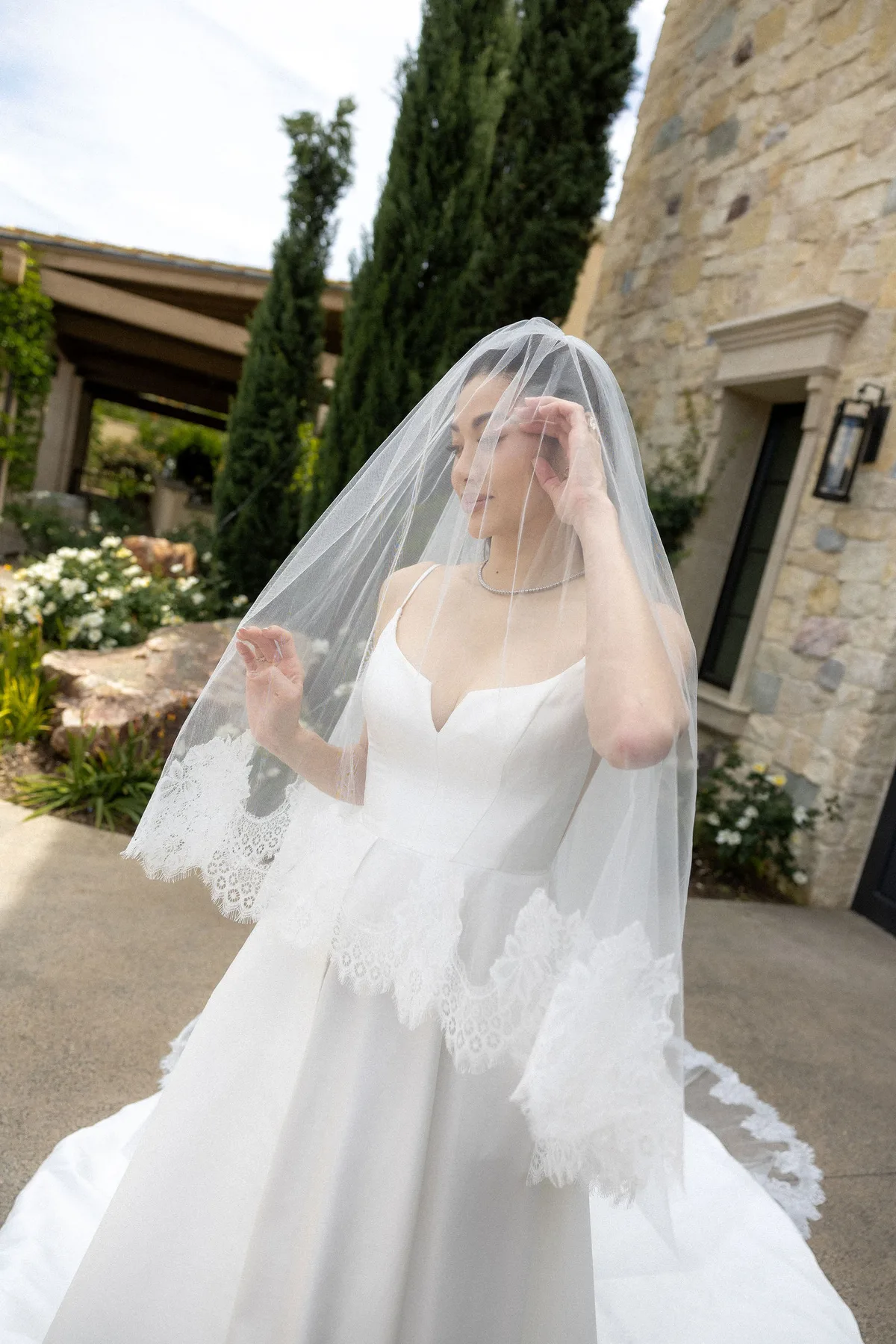 The bride lifting her veil on the path, cypress and stone behind