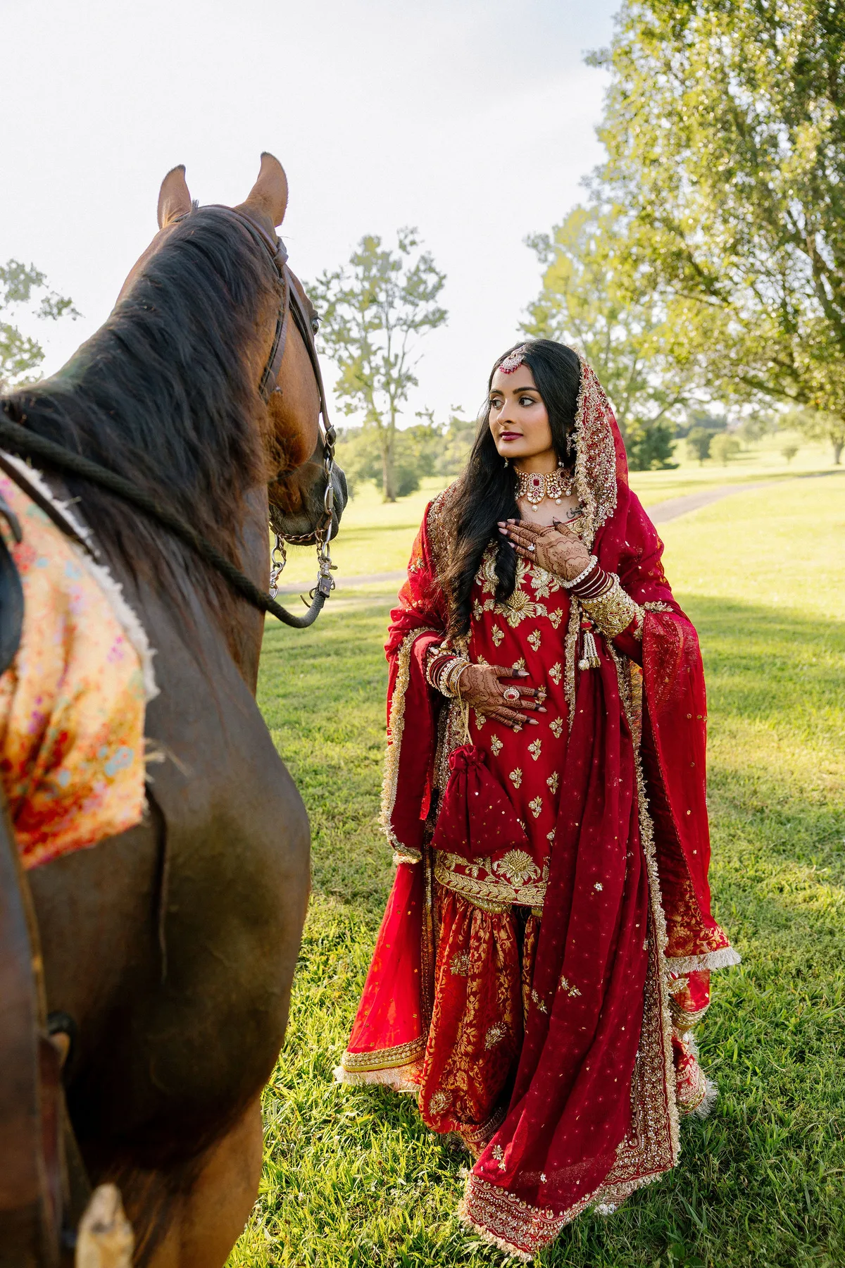 The bride in a red and gold lehenga beside a horse in the sun