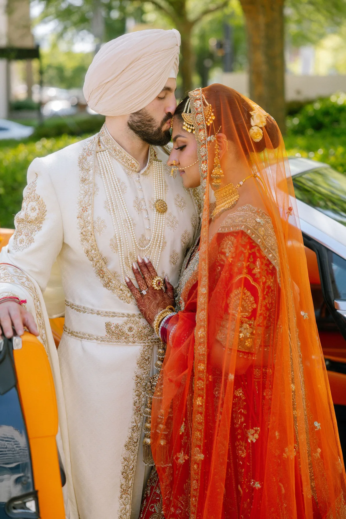The groom kissing the bride's forehead beside a car, her dupatta bright orange