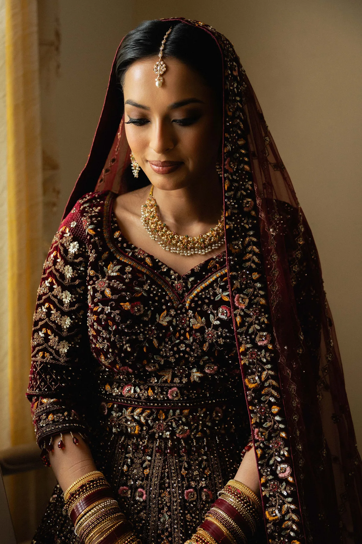 The bride in a deep red embroidered lehenga and gold jewellery, eyes lowered