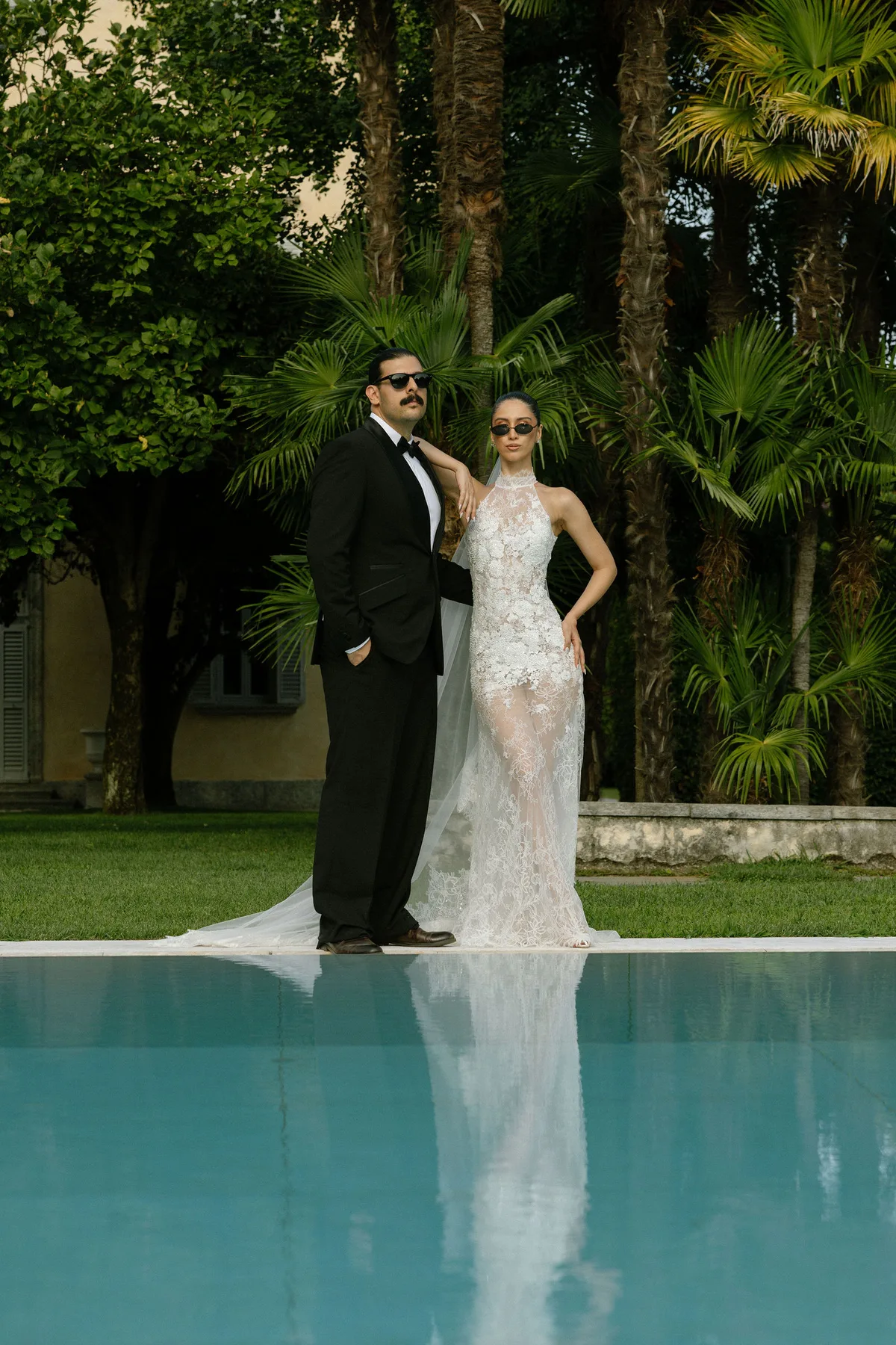 Bride and groom at the far end of a swimming pool, palms behind them