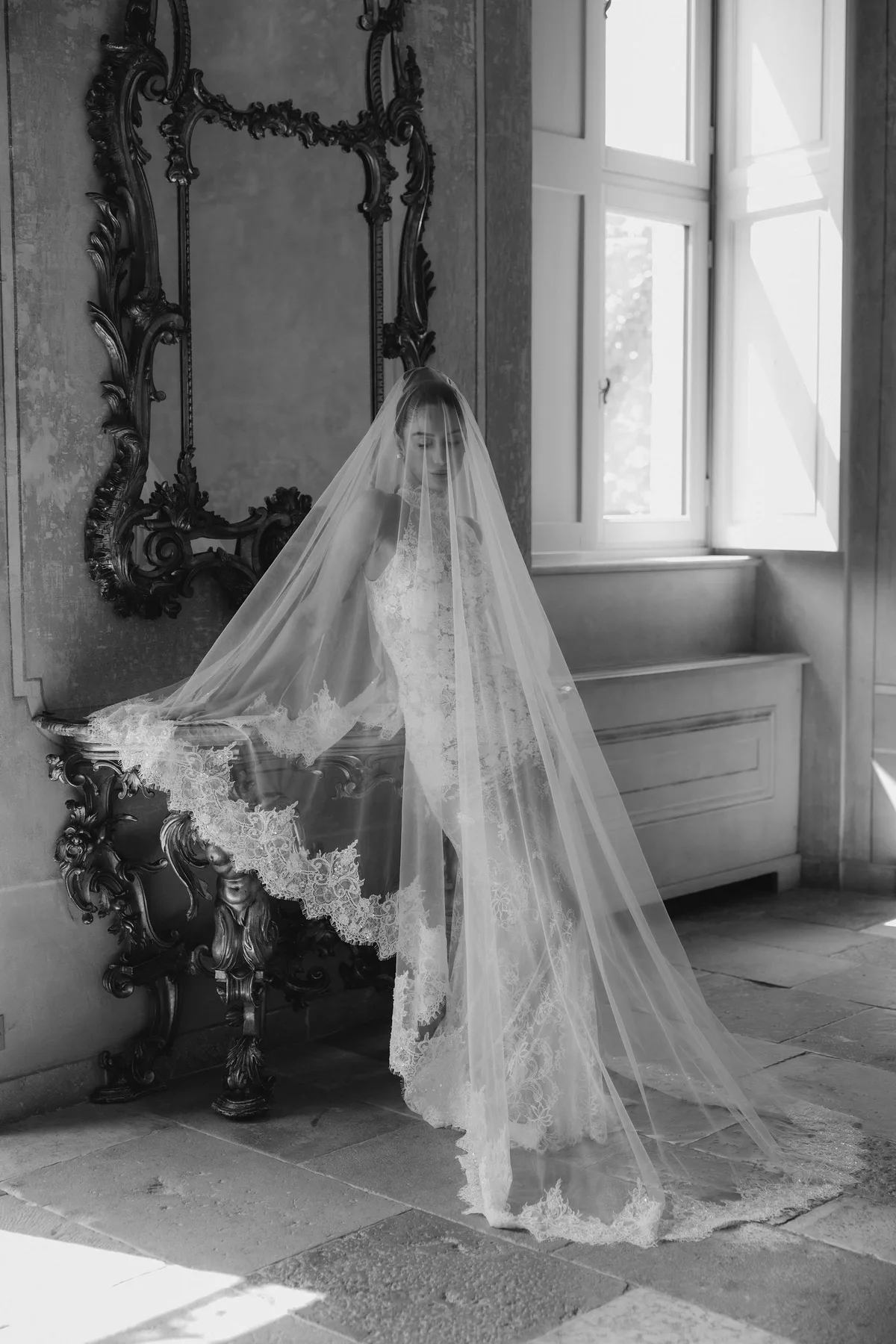 The bride by a tall window in a frescoed room, veil down, a carved mirror beside her