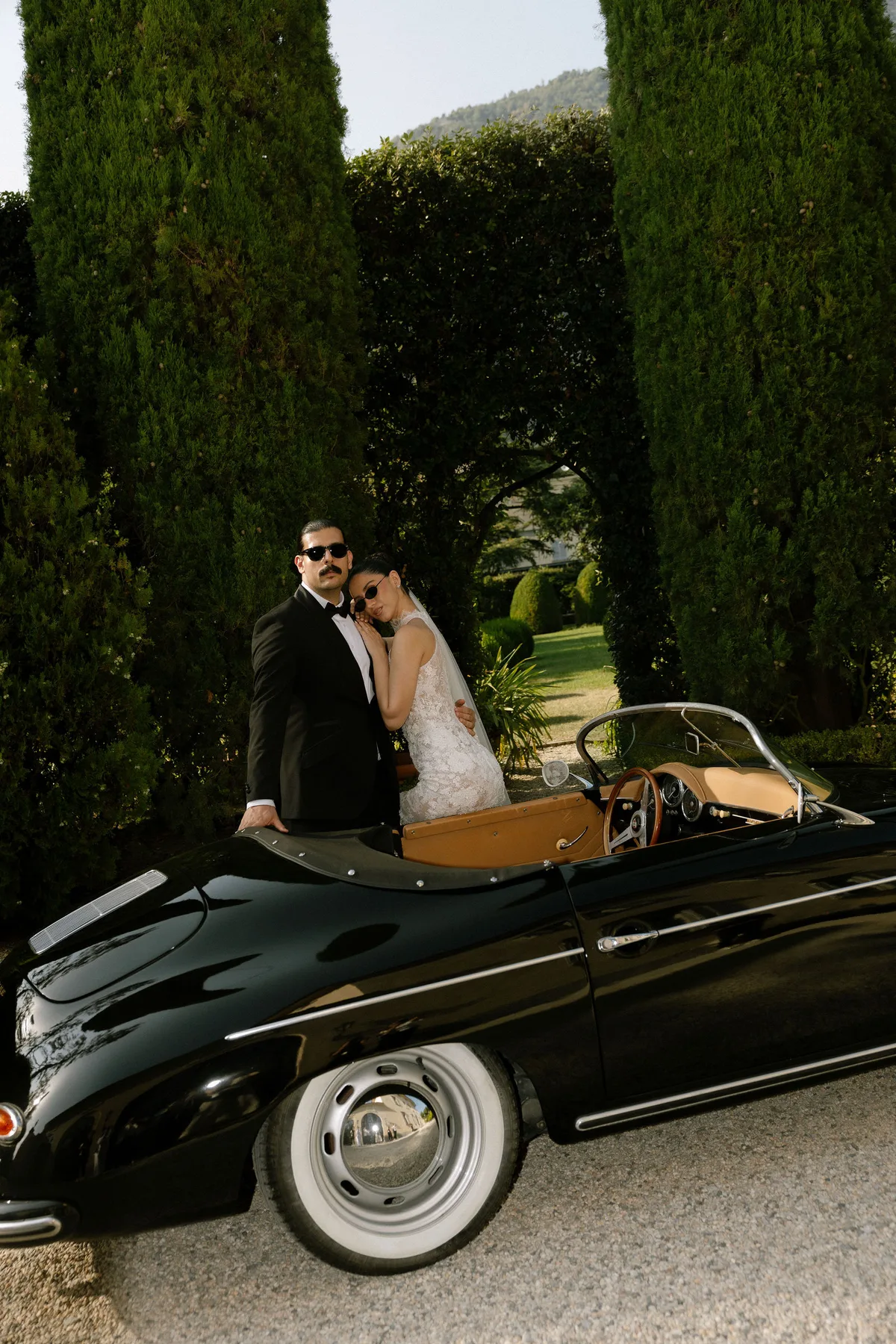 The couple leaning on an open black sports car between cypresses