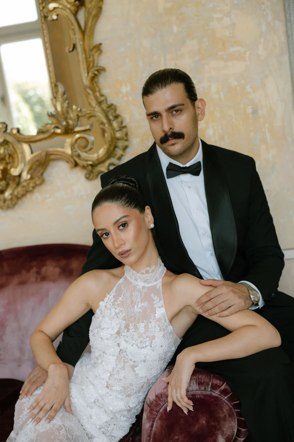The bride on a red velvet sofa, the groom's arms around her, a gilt mirror behind