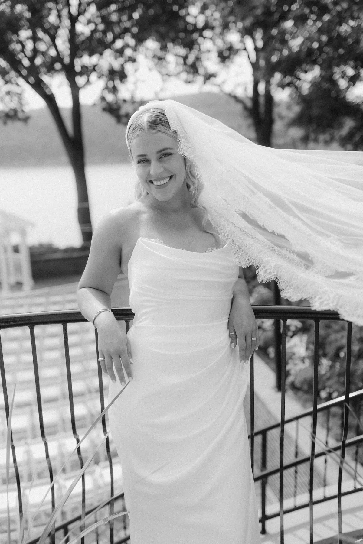 The bride laughing at an iron rail, her veil blown out behind, water beyond
